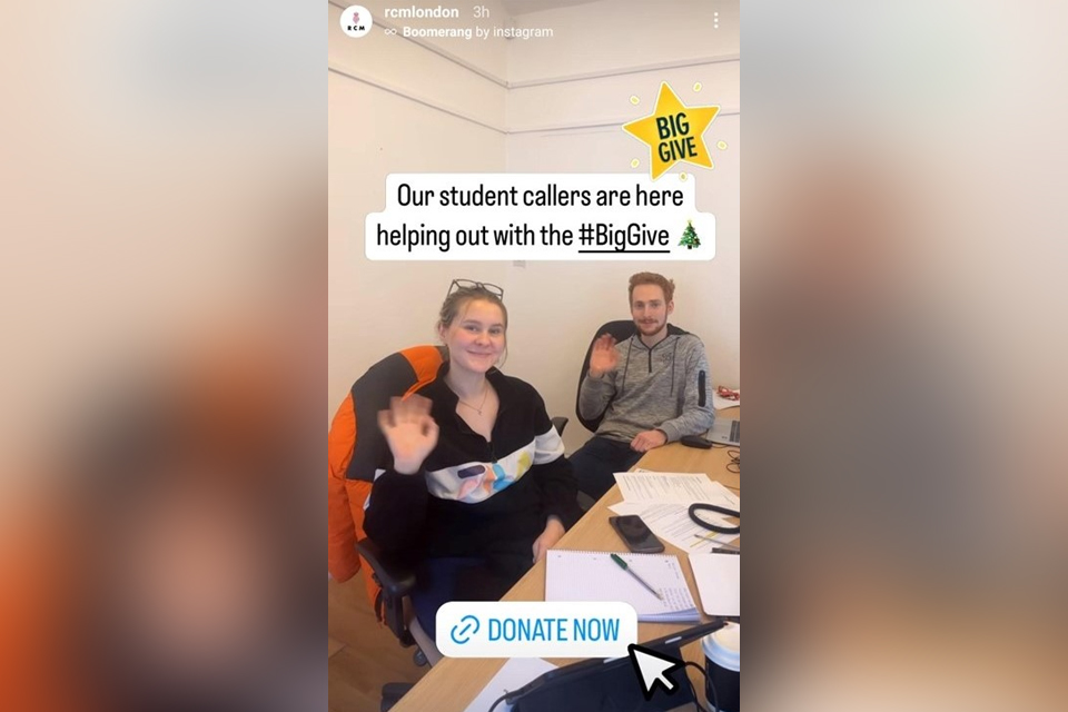 Two students waving at the camera, at a desk with text: Our student callers are here helping out with the Big Give